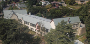 An aerial view of the Hillside Elementary School showing a somewhat meandering layout with a peaked roof and gables surrounded by pine trees.
