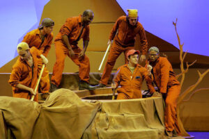 A staged production of Holes. The boys in the cast are in their orange jumpsuits, and Stanley is showing the others something he's found in his hole. The set is made from wooden supports covered with fabric.