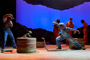 A staged production of Holes. Two actors are using puppets to represent a confrontation between a yellow-spotted lizard and a snake.