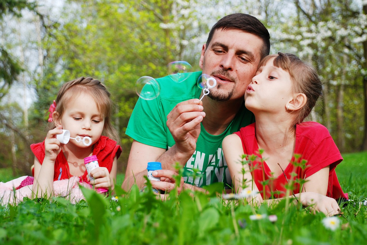 A father and daughters laying in the grass while blowing bubbles together.