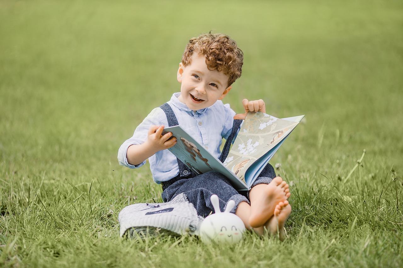 A small curly haired child sitting barefoot in the grass while reading a book.