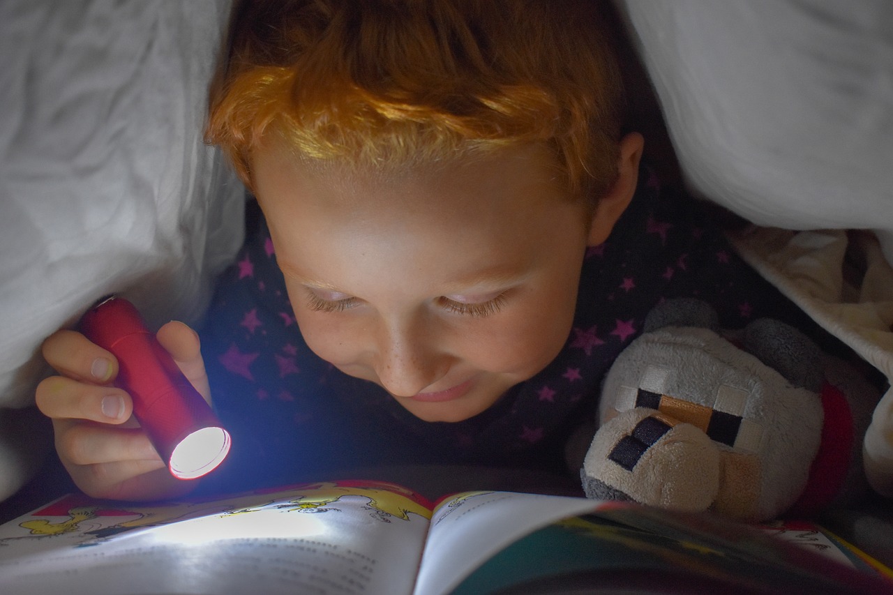 Small child reading a book by flashlight under a blanket.