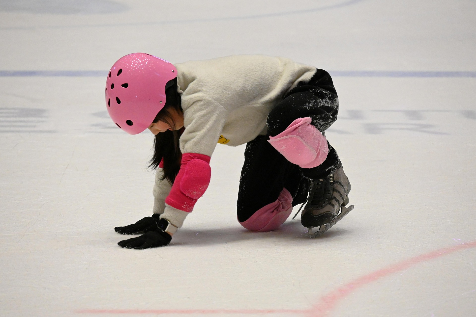 A child on the ice with knee and elbow pads, getting up while learning to skate.