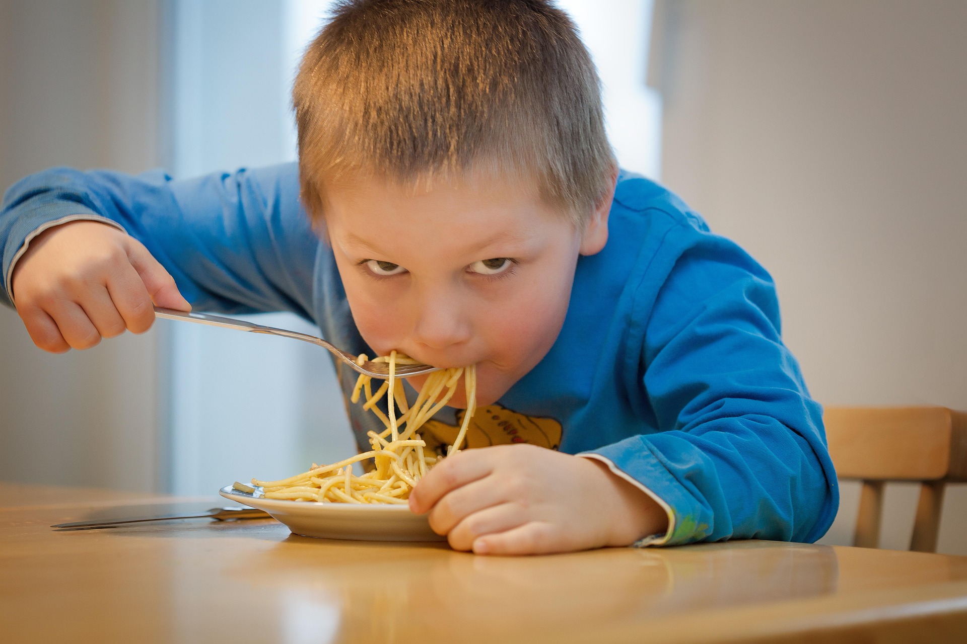 A child staring into the camera while putting noodles into their mouth.