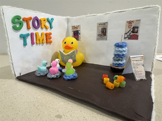 The winning entry of our Rubber Ducky Diorama Contest 2026. Created by Claire S., age 12. She's created a very cute story time scene, with a large rubber ducky reading a book to several smaller rubber duckies. The room has posters, play blocks, and knitted cushions for the kids to sit on.
